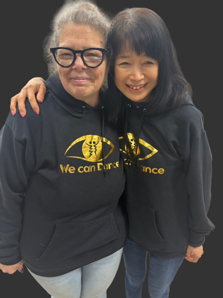 Two visually impaired participants, Rosa and Miyuki, smiling with their arms around each other while wearing matching black We Can Dance hoodies.