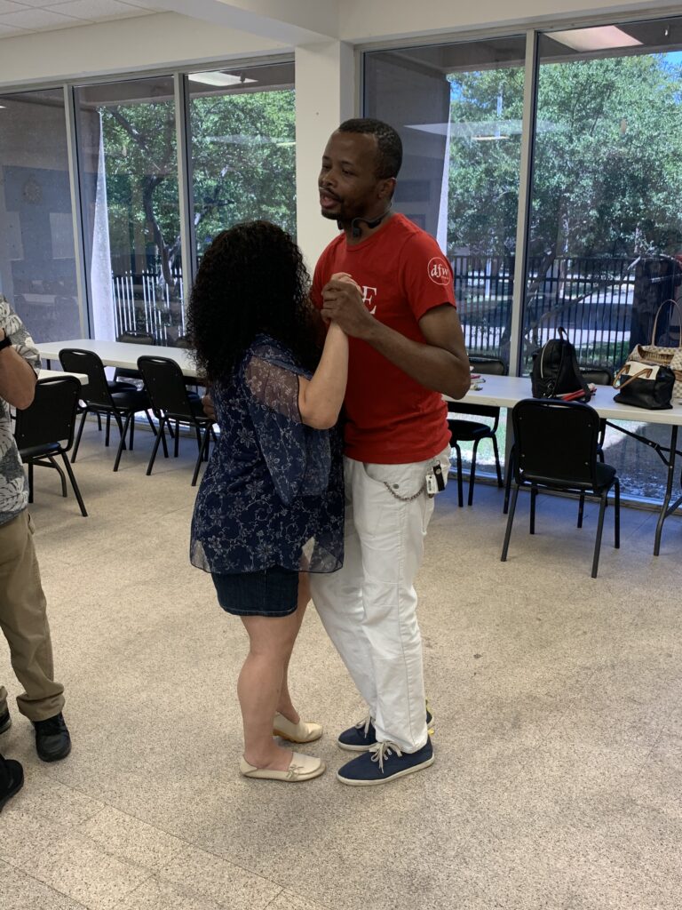 A participant in a red shirt practices partner dancing with a woman during a Dance in the Dark workshop.