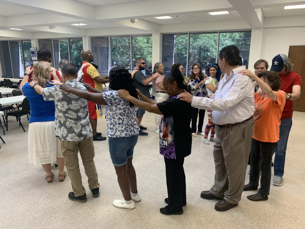 Participants form a connected circle with their hands on one another’s shoulders during a group dance activity.